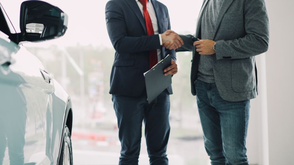 Dois homens em roupas formais trocando um aperto de mão ao lado de um carro, simbolizando uma negociação ou acordo de negócios no setor automotivo.