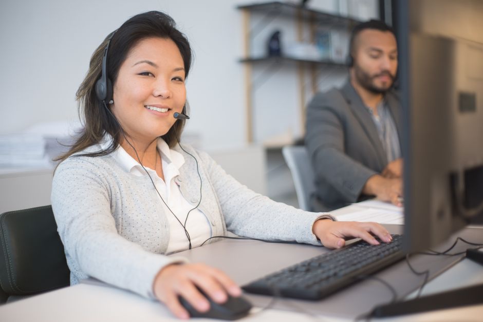 Profissional de suporte técnico ou atendimento ao cliente sorrindo enquanto trabalha em um computador em um ambiente de escritório moderno.