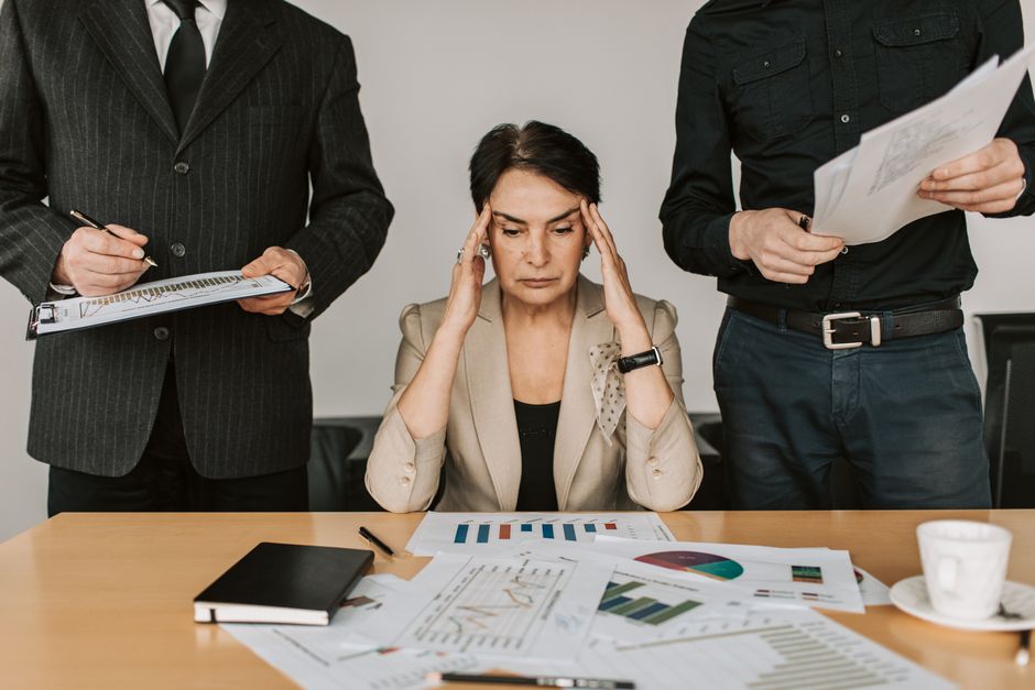 Mulher estressada lidando com pressão financeira ou problemas de gestão enquanto três colegas consultam documentos ao seu redor.
