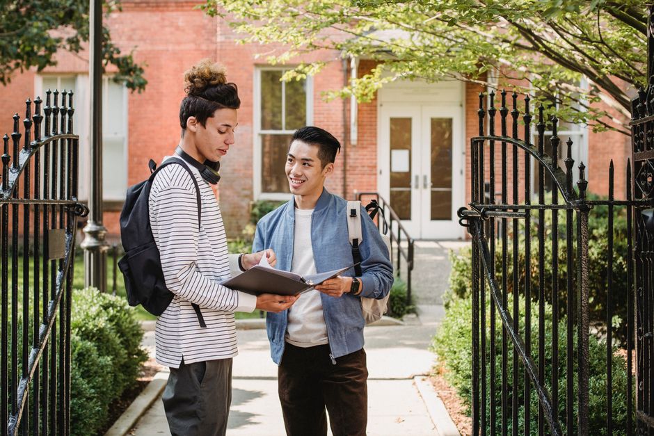 Dois estudantes conversando ao ar livre, possivelmente discutindo ideias acadêmicas ou projetos, em frente a uma casa com jardins bem cuidados.