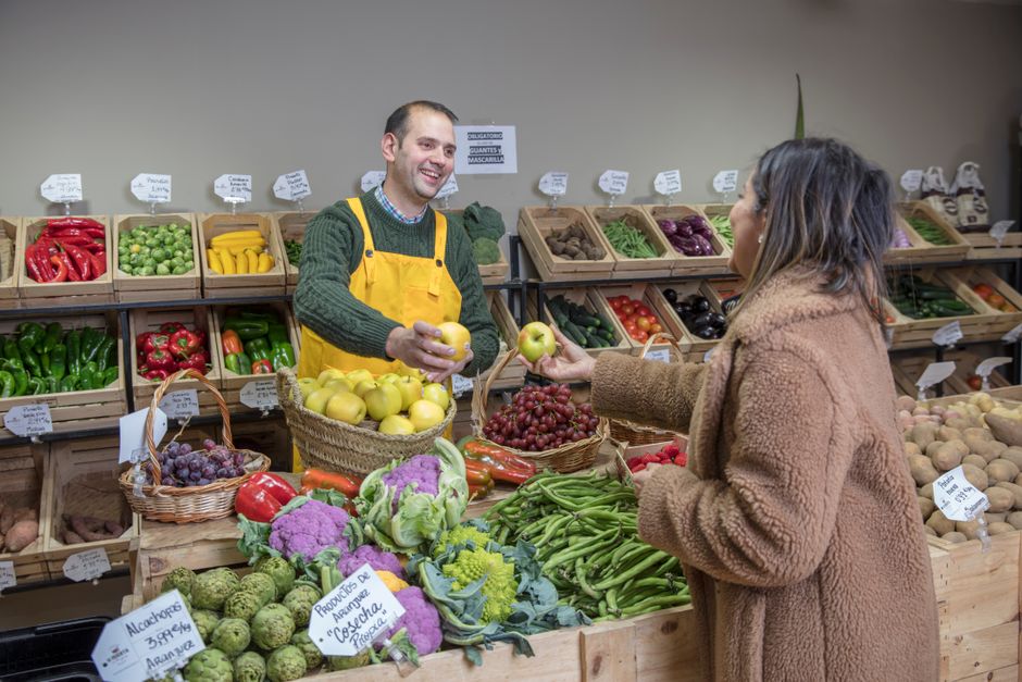Cliente comprando frutas e verduras em feira orgânica, recebendo atendimento do vendedor sorridente.