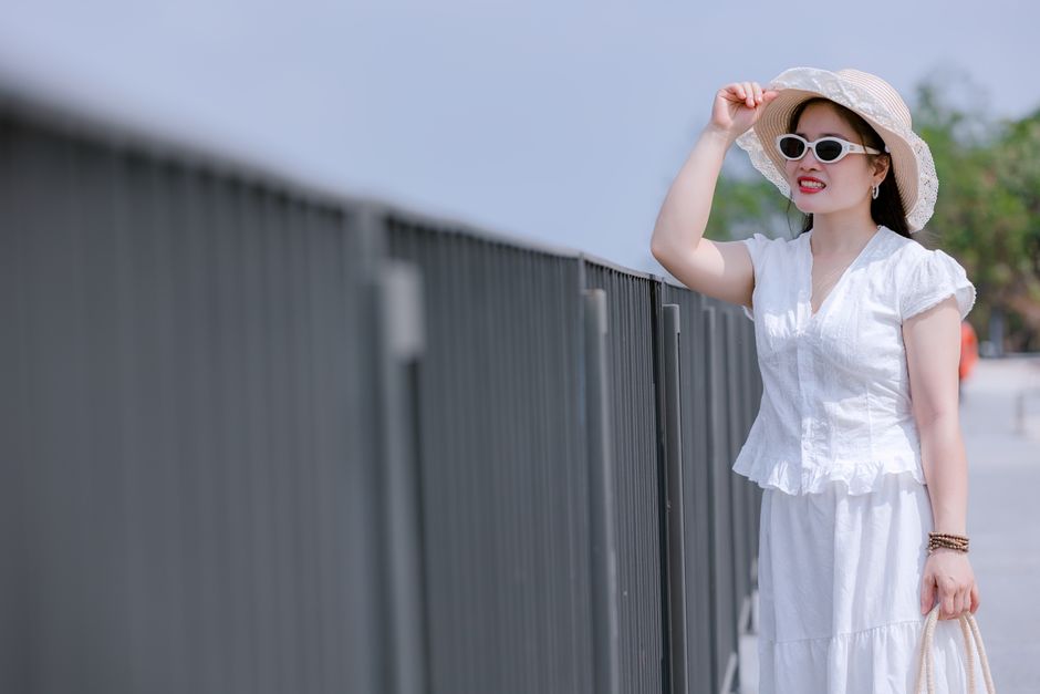 Mulher vestida de branco com chapéu e óculos, apreciando o céu ao ar livre em um ambiente ensolarado.