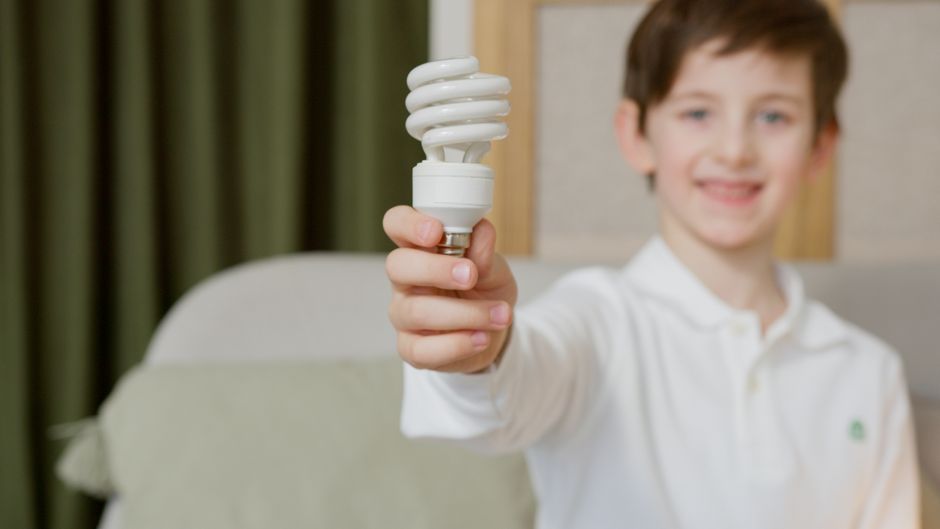Menino sorridente segurando uma lâmpada de luz fluorescente compacta, representando ideias e inovação.