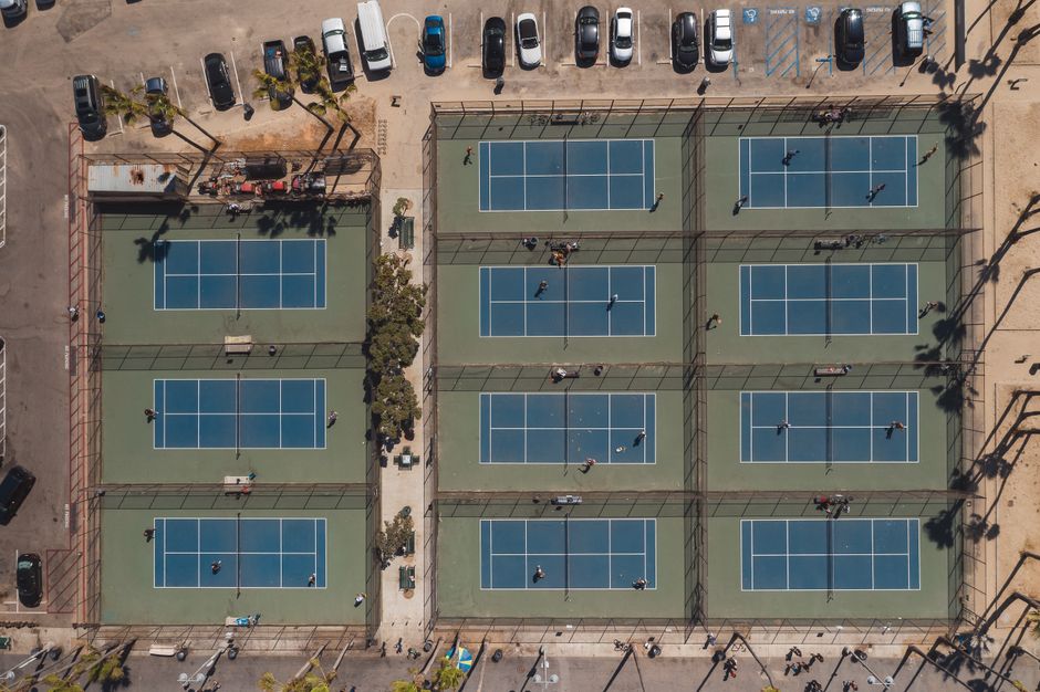 Visão aérea de quadras de tênis em um espaço ao ar livre, com pessoas praticando o esporte em um dia ensolarado.
