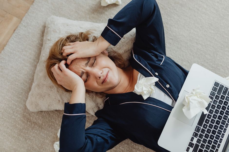 Mulher descansando no chão com expressão de cansaço ao lado de um laptop, representando estresse ou fadiga no ambiente de trabalho ou estudos.