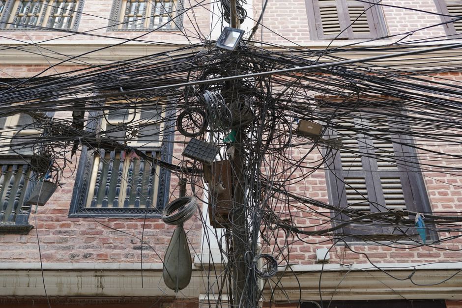Imagem de um poste elétrico com cabos desordenados em frente a um edifício residencial, ilustrando problemas de infraestrutura urbana.