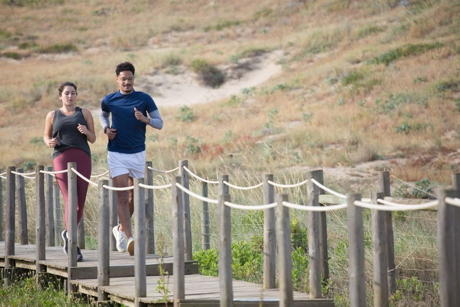 Homens praticando corrida ao ar livre em ambiente natural com trilha de madeira e vegetação ao redor.