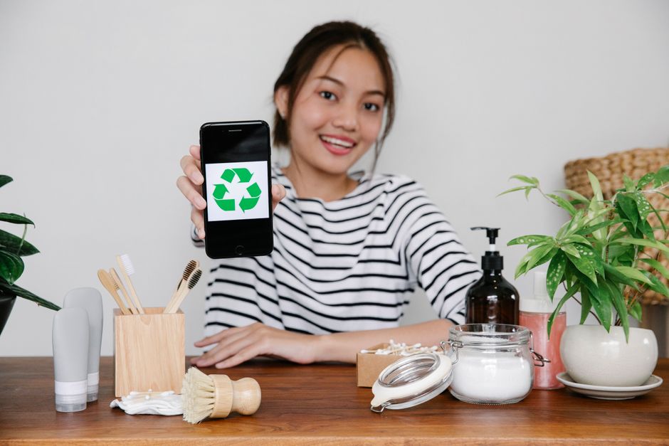 Mulher sorridente exibindo um celular com símbolo de reciclagem, promovendo a importância da sustentabilidade e reciclagem no contexto do cuidado ambiental.