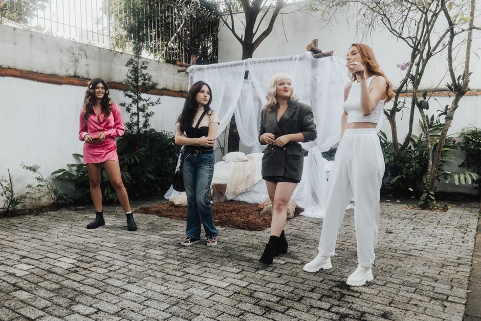 Grupo de jovens em uma sessão de fotos ao ar livre, com cenário decorativo de paredes brancas, plantas e tecidos sustentando uma atmosfera moderna e descontraída.