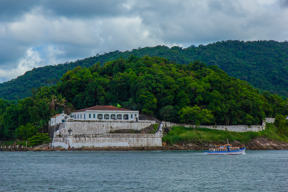 Vista de uma fortaleza histórica localizada à beira-mar, cercada por vegetação exuberante, com um barco navegando na água próxima, representando patrimônio cultural e turismo costeiro.