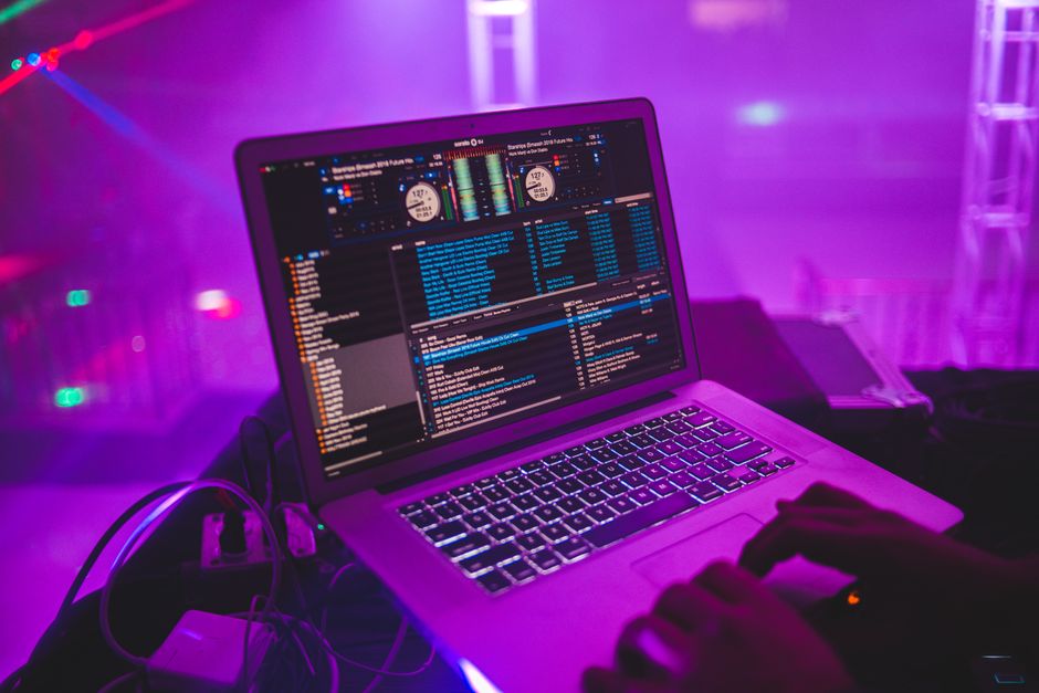 Pessoa operando um computador com software de leitura de músicas em um ambiente com iluminação colorida, possivelmente durante um evento de tecnologia ou DJ ao vivo.