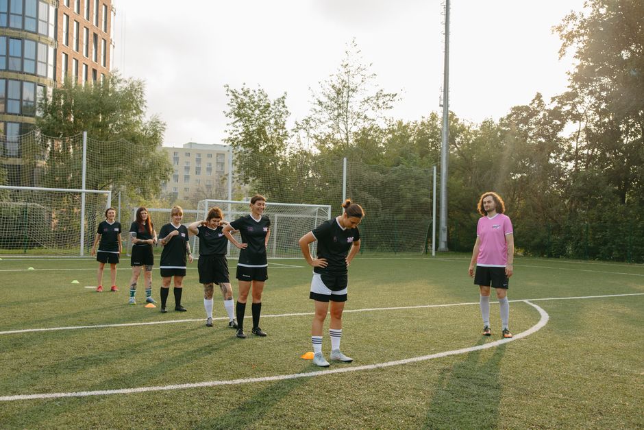 Um grupo de jogadoras de futebol feminino se preparando para treinamento ou jogo em um campo ao ar livre, com árvores e edifícios ao fundo, ao pôr do sol.