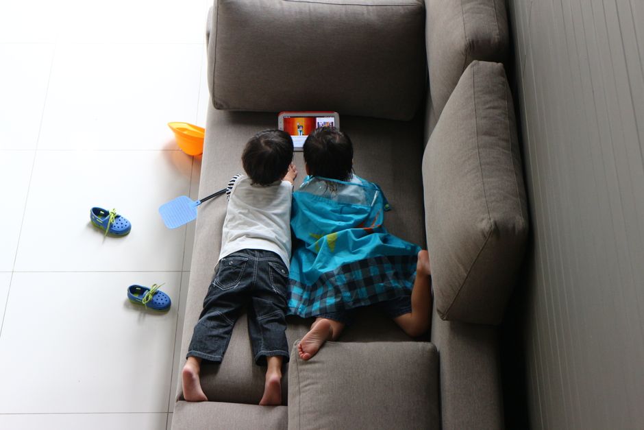 Crianças assistindo a um conteúdo em um tablet deitados no sofá, refletindo o uso de tecnologia e entretenimento infantil em casa.