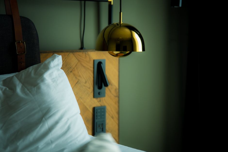 Imagem de uma cabeceira de cama com uma luminária dourada moderna ao lado, refletindo um ambiente acolhedor e contemporâneo, ideal para artigos de decoração ou hotelaria.