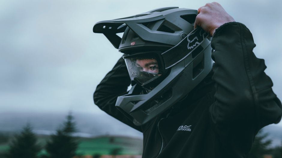 Pessoa ajustando o capacete de proteção para atividades ao ar livre ou esportes motorizados, em ambiente ao ar livre com céu nublado e vegetação ao fundo.