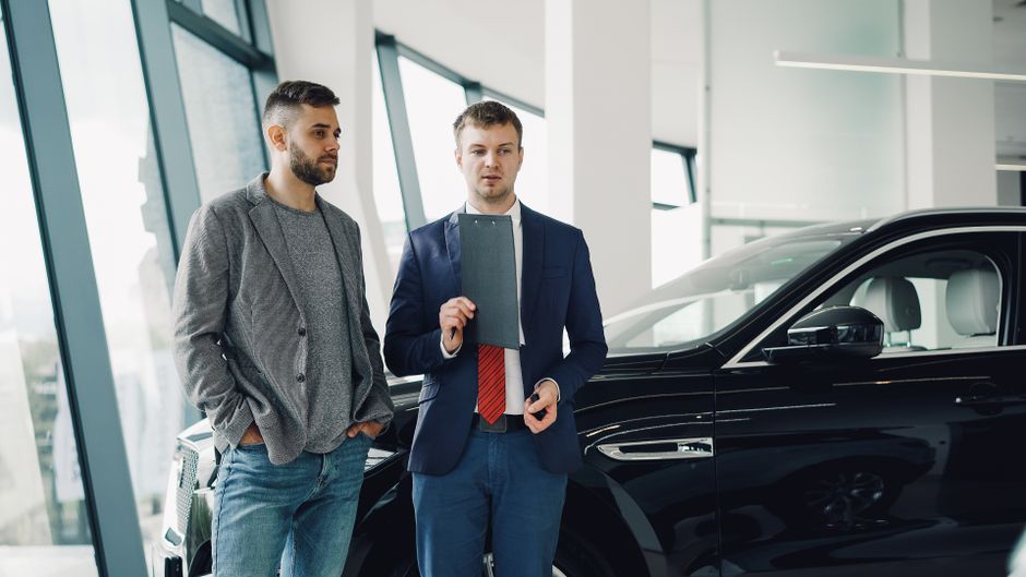 Dois homens em uma concessionária de carros discutem opções de compra ou venda de veículos em um ambiente de negócios.