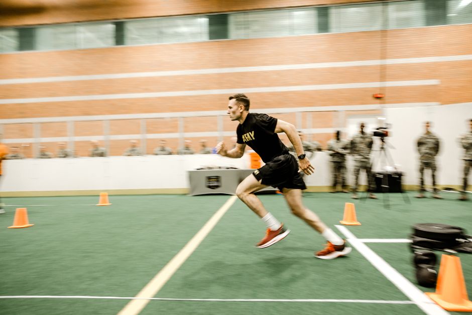 Homem treinando corrida de velocidade em ambiente indoor com obstáculos e pessoas acompanhando ao fundo, destacando foco em preparação física e desempenho esportivo.