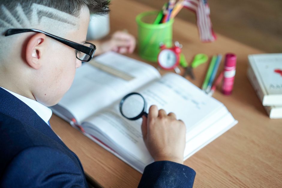 Garoto estudando com foco, usando lupa para leitura, em ambiente escolar ou de estudos, com materiais escolares ao redor.