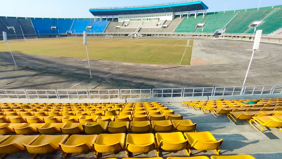 Estádio de futebol vazio com arquibancadas amarelas em uma manhã ensolarada, pronto para receber uma partida ou evento esportivo.