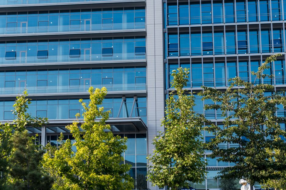 Fachada de prédio de escritórios contemporâneo com grande quantidade de janelas de vidro refletindo o céu, cercado por árvores verdes.