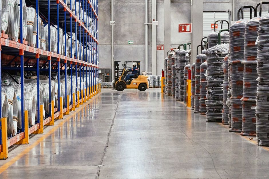 Imagem de um armazém industrial com prateleiras cheias de materiais e um operador de empilhadeira ao fundo, evidenciando logística e armazenamento empresarial.
