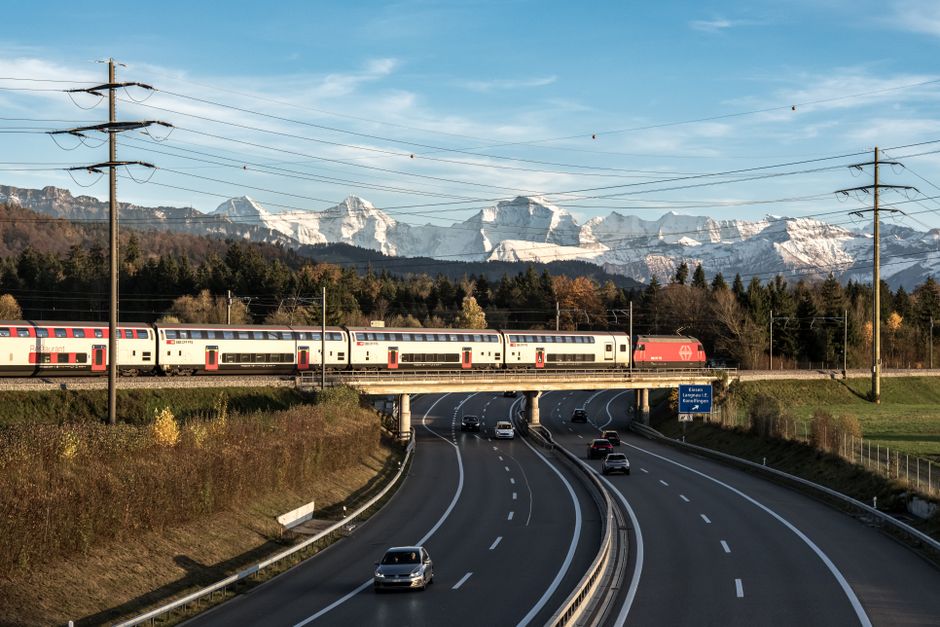 Imagem de uma ferrovia passando sobre uma estrada, com montanhas ao fundo, representando mobilidade e infraestrutura de transporte.