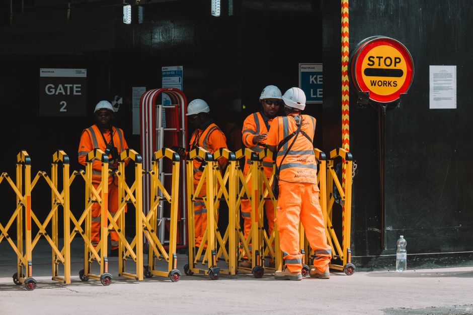 Grupo de trabalhadores em direção a uma área de segurança na construção civil, com sinalização de parada de obras ao fundo.