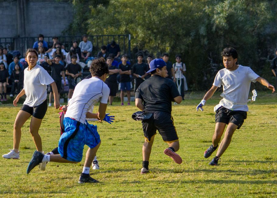 Grupo de jovens jogando uma partida de futebol ao ar livre, com espectadores ao fundo, em um ambiente verde e ensolarado.