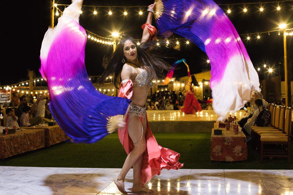 Dançarina realizando apresentação cultural com véu colorido em evento ao ar livre à noite, sob iluminação de luzes decorativas.