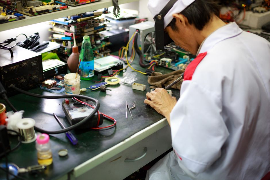 Profissional reparando componentes eletrônicos em um ambiente de oficina com diversas ferramentas e peças ao redor.
