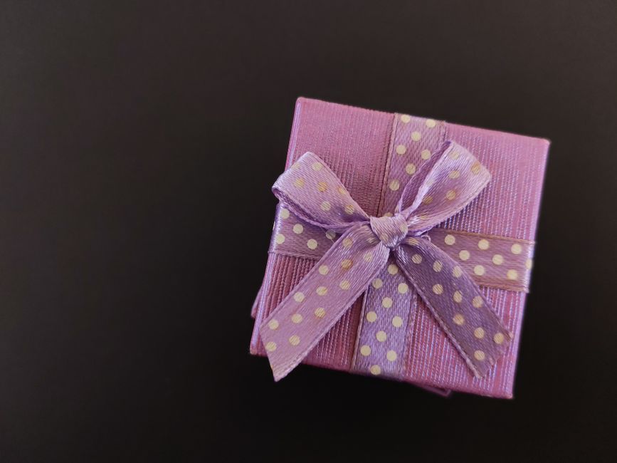 Imagem de uma caixa de presente rosa decorada com fita de cetim com bolinhas douradas, sobre fundo escuro.