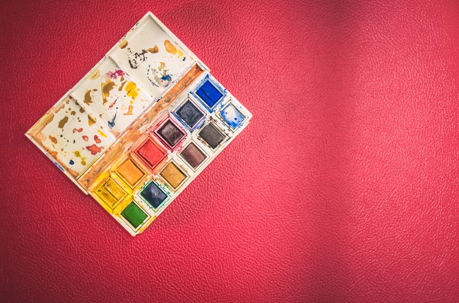 Paleta de tintas aquarela com cores vibrantes sobre fundo vermelho texturizado, usada como ilustração de criatividade artística ou técnicas de pintura.