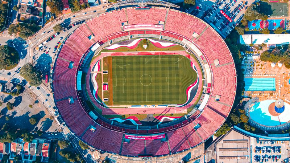Imagem aérea de um estádio de futebol com arquibancadas coloridas, visto de cima, destacando a estrutura e o campo central.