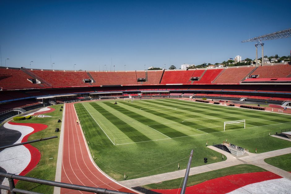 Imagem de um estádio de futebol vazio, pronto para uma partida, com arquibancadas e campo bem preservados sob um céu claro.