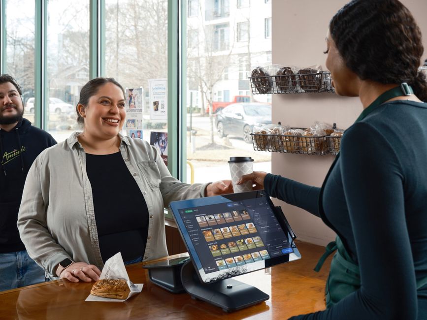 Mulher fazendo pagamento com cartão de débito em estabelecimento de cafeteria, com atendente e cliente sorridente ao fundo, em ambiente de comércio ao estilo de uma padaria ou café.