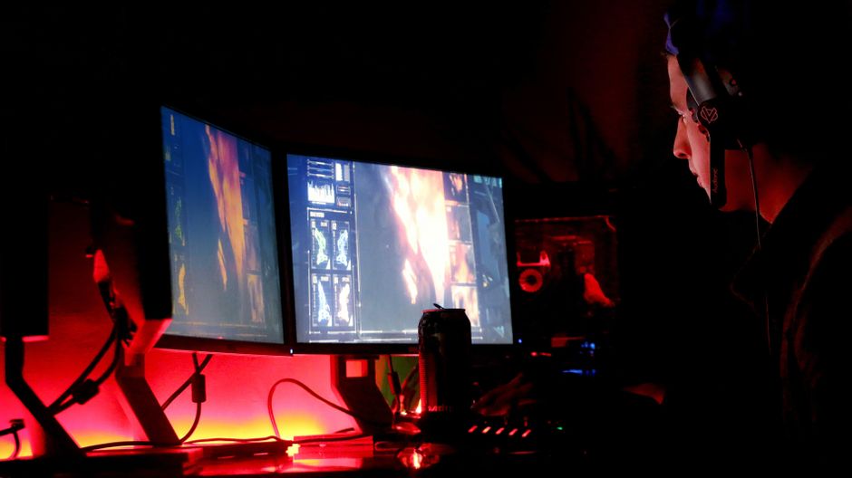 Pessoa jogando videogame em ambiente escuro com duas telas e iluminação ambiente vermelha, destacando o universo dos jogos eletrônicos.