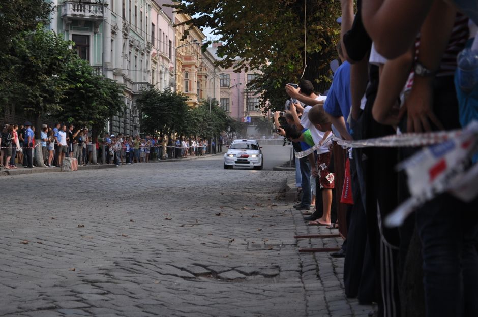 Mulheres e crianças assistindo a uma corrida de carros em uma rua urbana, com pessoas usando celulares para registrar o momento.