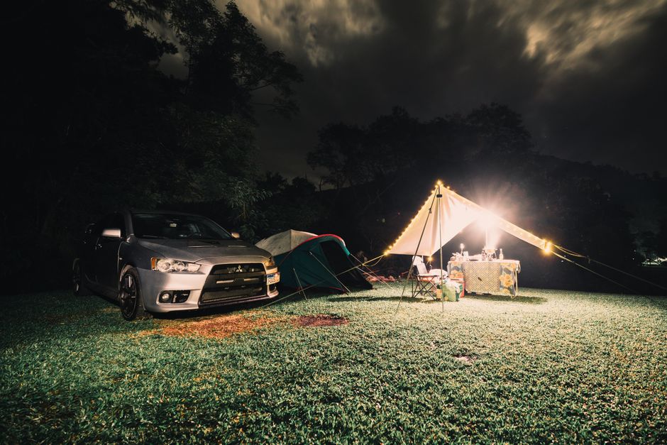 Cena de acampamento ao ar livre à noite, com uma caminhonete, uma tenda, uma mesa com comida e iluminação decorativa, criando uma atmosfera aconchegante na natureza.