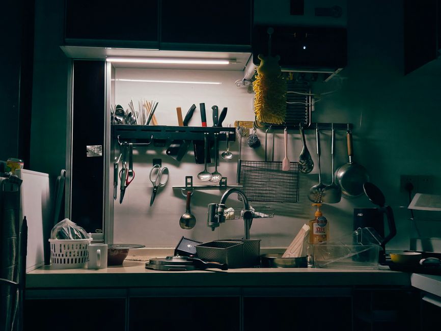 Imagem de uma cozinha com iluminação de luz fria, contendo utensílios de cozinha pendurados e organização de itens domésticos.