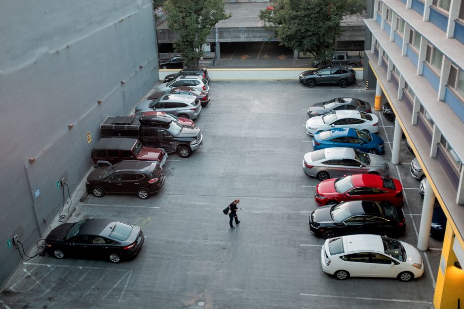 Imagem de um estacionamento coberto com várias vagas ocupadas e uma pessoa caminhando ao centro, vista de um prédio alto em ambiente urbano.