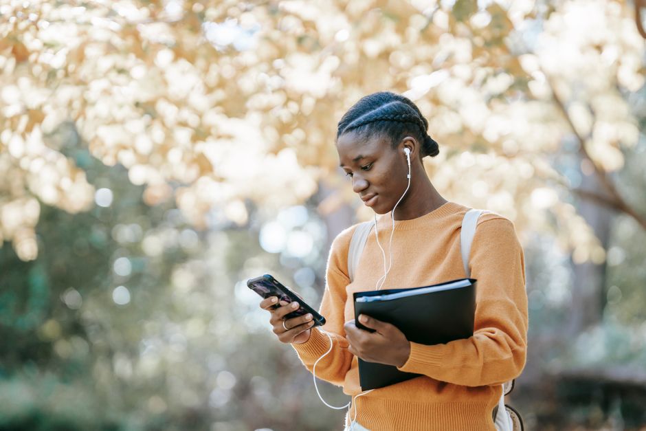Mulher jovem usando celular e fones de ouvido ao ar livre, com expressiva presença de elementos naturais ao fundo, representando conexão digital e convivência com a natureza.