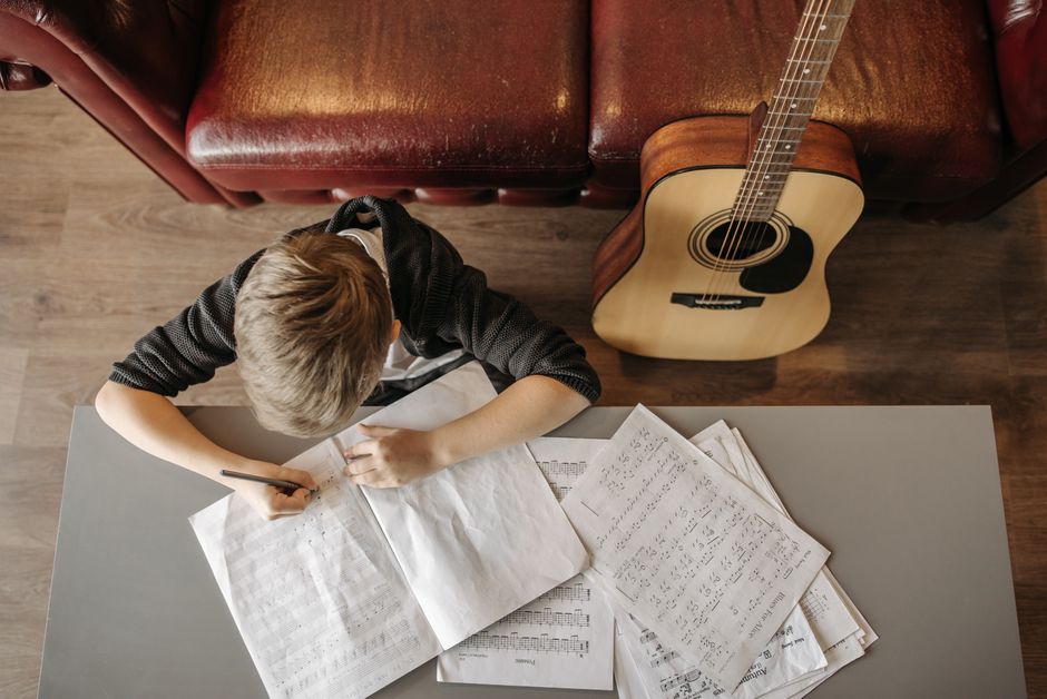Jovem estudando e fazendo anotações enquanto trabalha com música ao fundo, com uma guitarra ao lado e uma sala de estar confortável.
