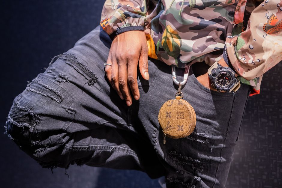 Imagem de uma pessoa usando roupa casual, destacando acessórios como relógio e chaveiro de marca de luxo, sugerindo estilo e personalidade urbana.