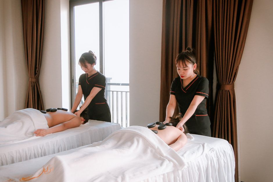 Duas terapeutas realizando sessões de massagem em duas pessoas em um ambiente de spa relaxante com iluminação natural.