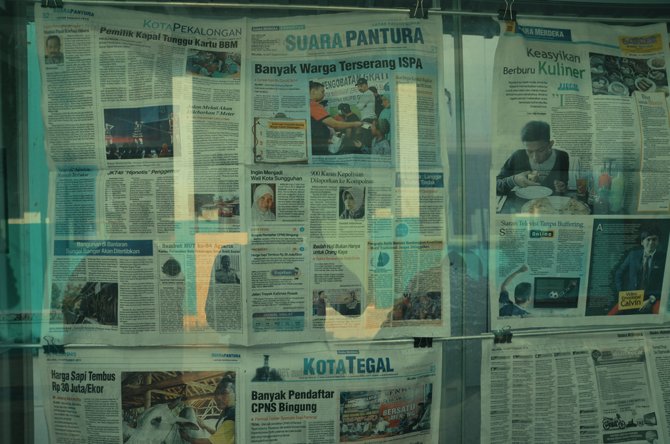 Jornal impresso em uma banca de jornal exibindo notícias diversas, com uma pessoa lendo enquanto come, refletindo uma rotina urbana de consumo de notícias.