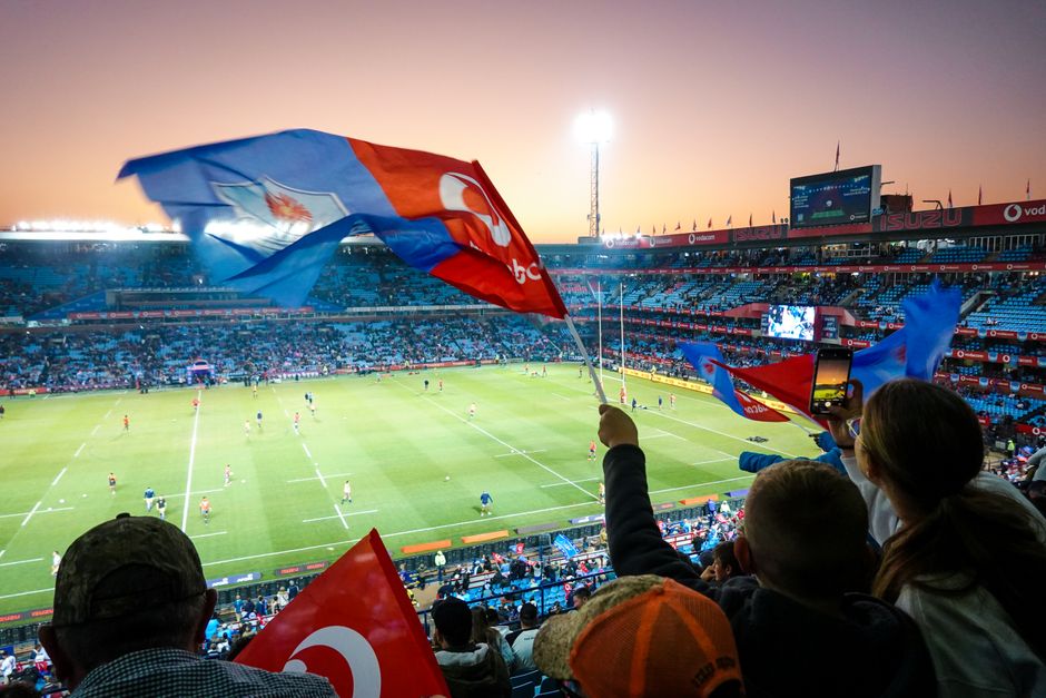 Torcida assistindo a uma partida de futebol em estádio ao entardecer, com bandeiras e clima de entusiasmo.
