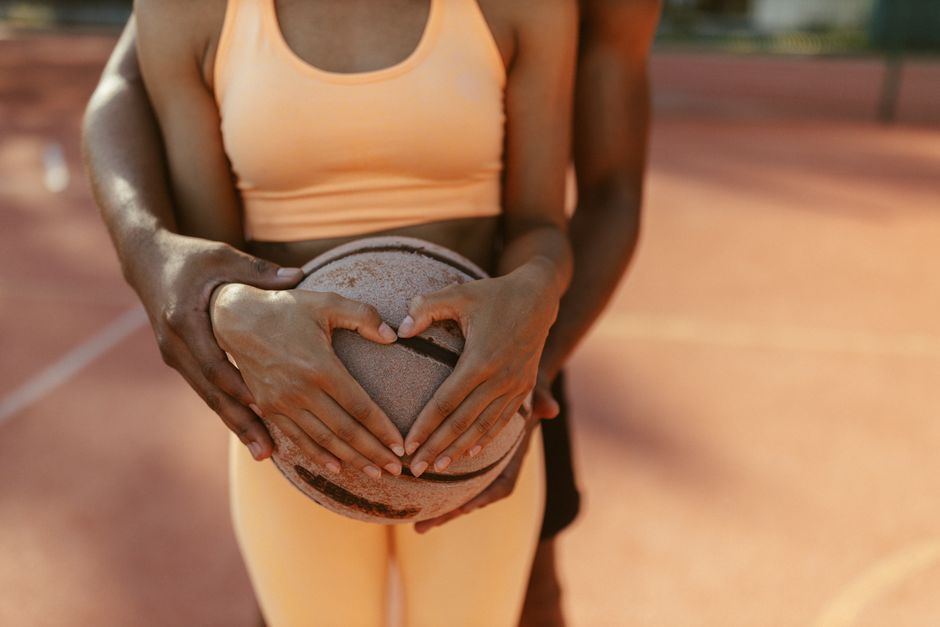 Duas jogadoras de basquete formando um coração com as mãos enquanto seguram uma bola em uma quadra ao ar livre, simbolizando união e paixão pelo esporte.