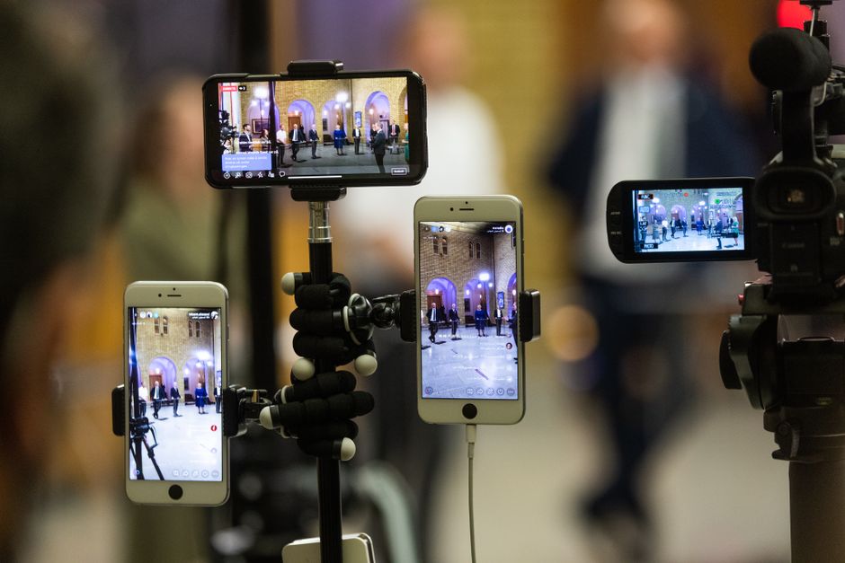 Imagem de profissionais de mídia capturando uma apresentação ou evento com vários smartphones e câmeras em um ambiente interno.