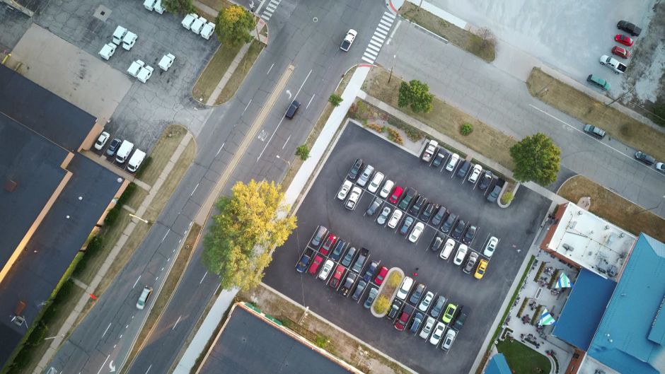 Imagem de vista aérea mostrando um estacionamento com carros organizados, em área urbana com ruas e edifícios ao redor, ideal para ilustrar temas relacionados a mobilidade urbana ou planejamento de tráfego.