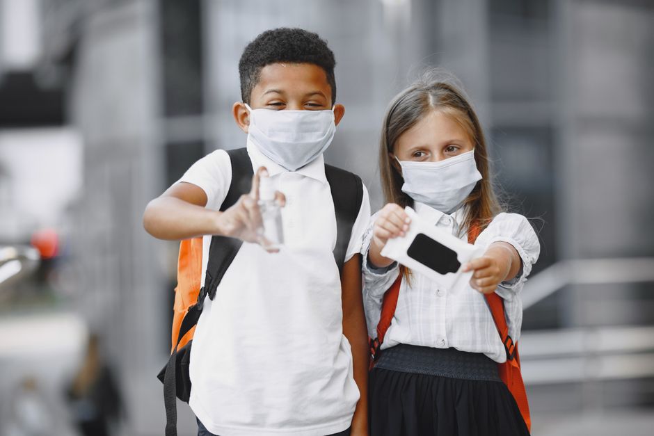 Duas crianças usando máscara facial, dando um aperto de mão enquanto estão na rua com mochilas, sugerindo tema de retorno às aulas ou convivência segura em contextos escolares.